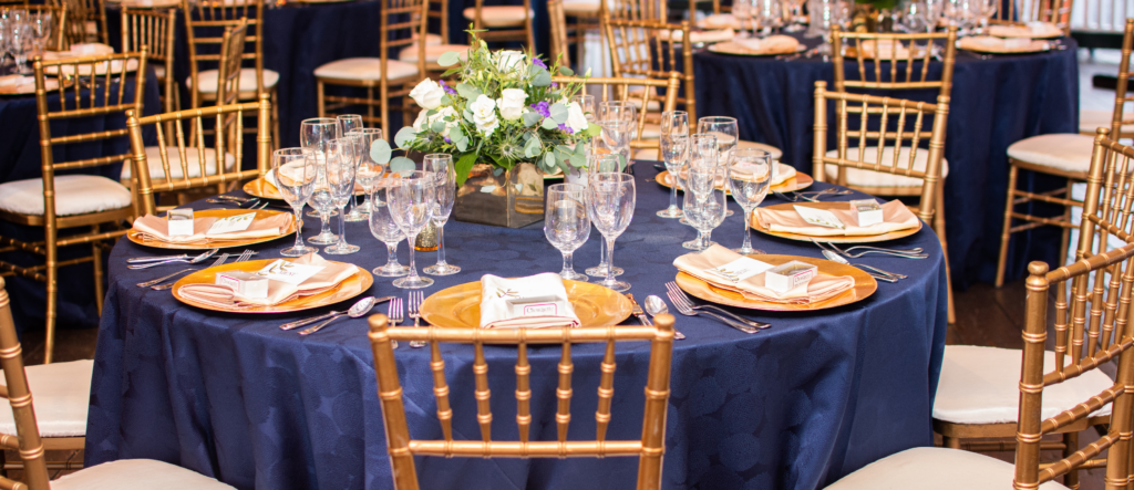 Elegant gala table setting featuring royal blue tablecloths, gold Chiavari chairs, and stunning floral centerpieces